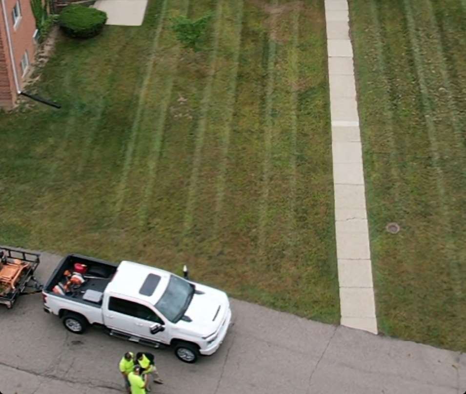 Drone view of a white pickup with a mower trailer and three crew members in hi-vis shirts beside a freshly striped apartment lawn