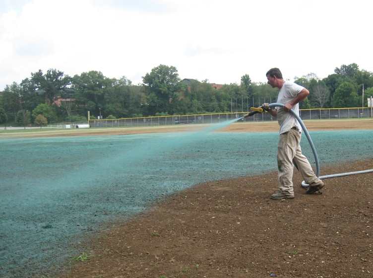 A worker spraying green hydroseed mulch across a bare ball field