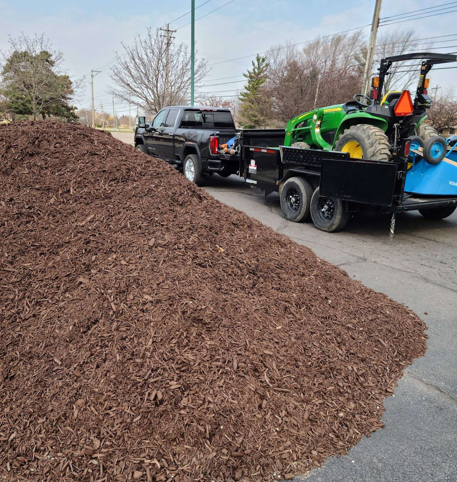 A large mulch pile beside a pickup and a trailer carrying landscape equipment