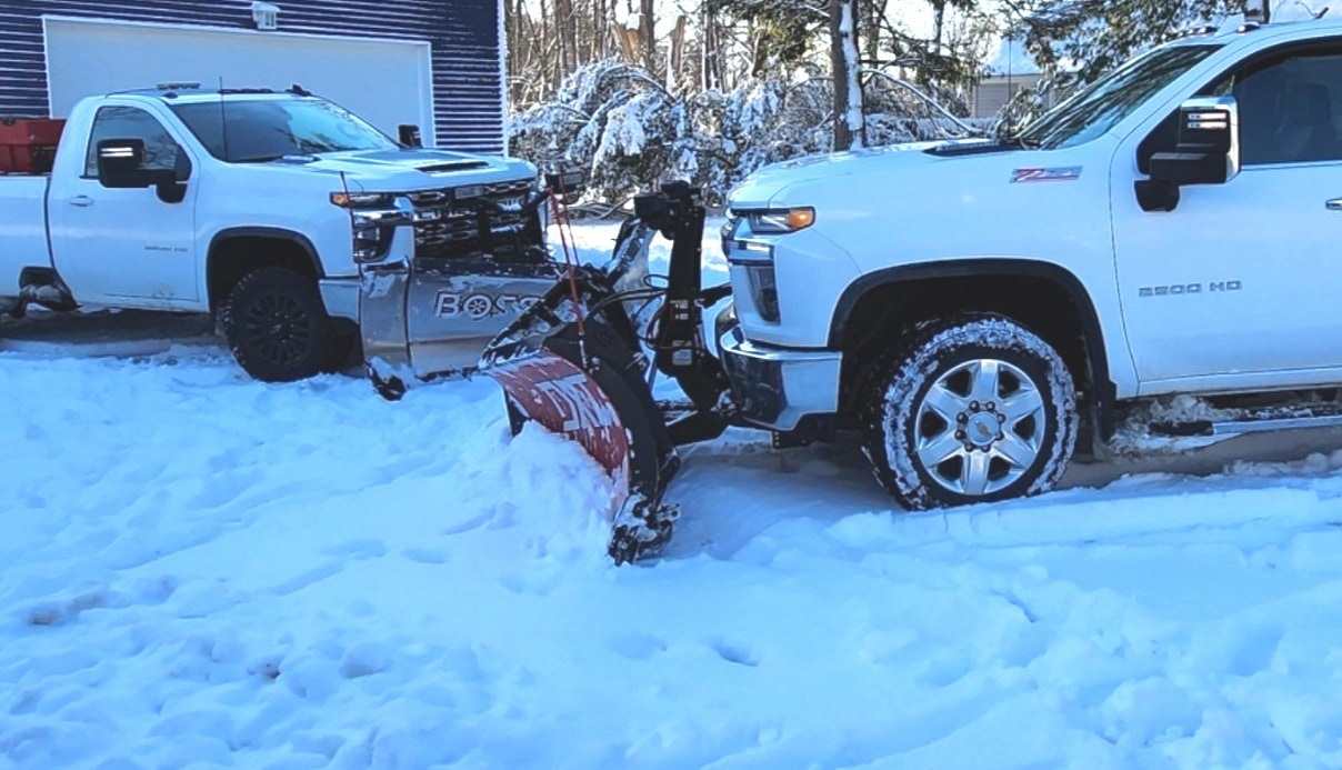 Two white Chevrolet heavy-duty pickups with V-plows in fresh snow